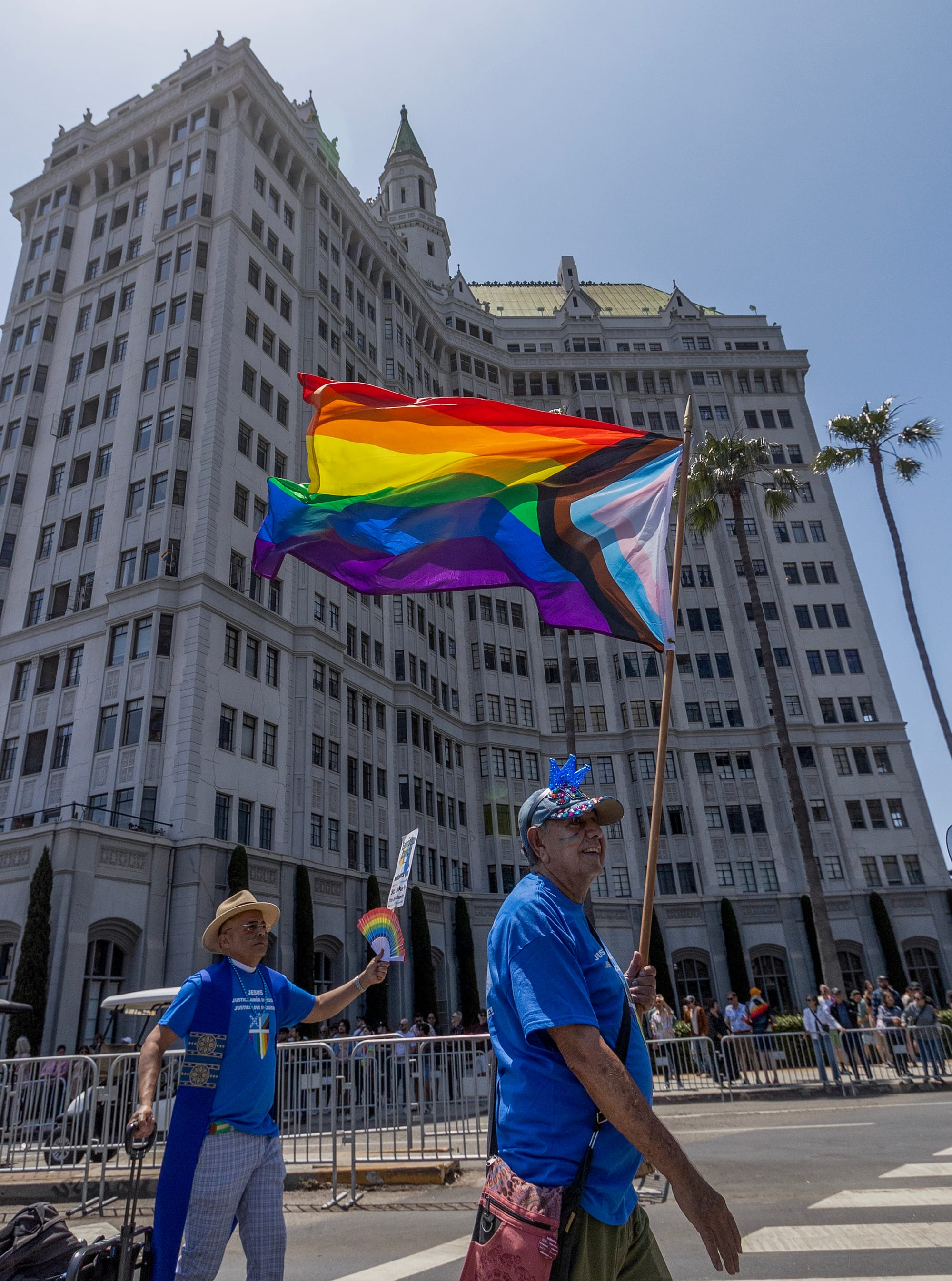A man carries a rainbow-colored flag in front of a tall building.