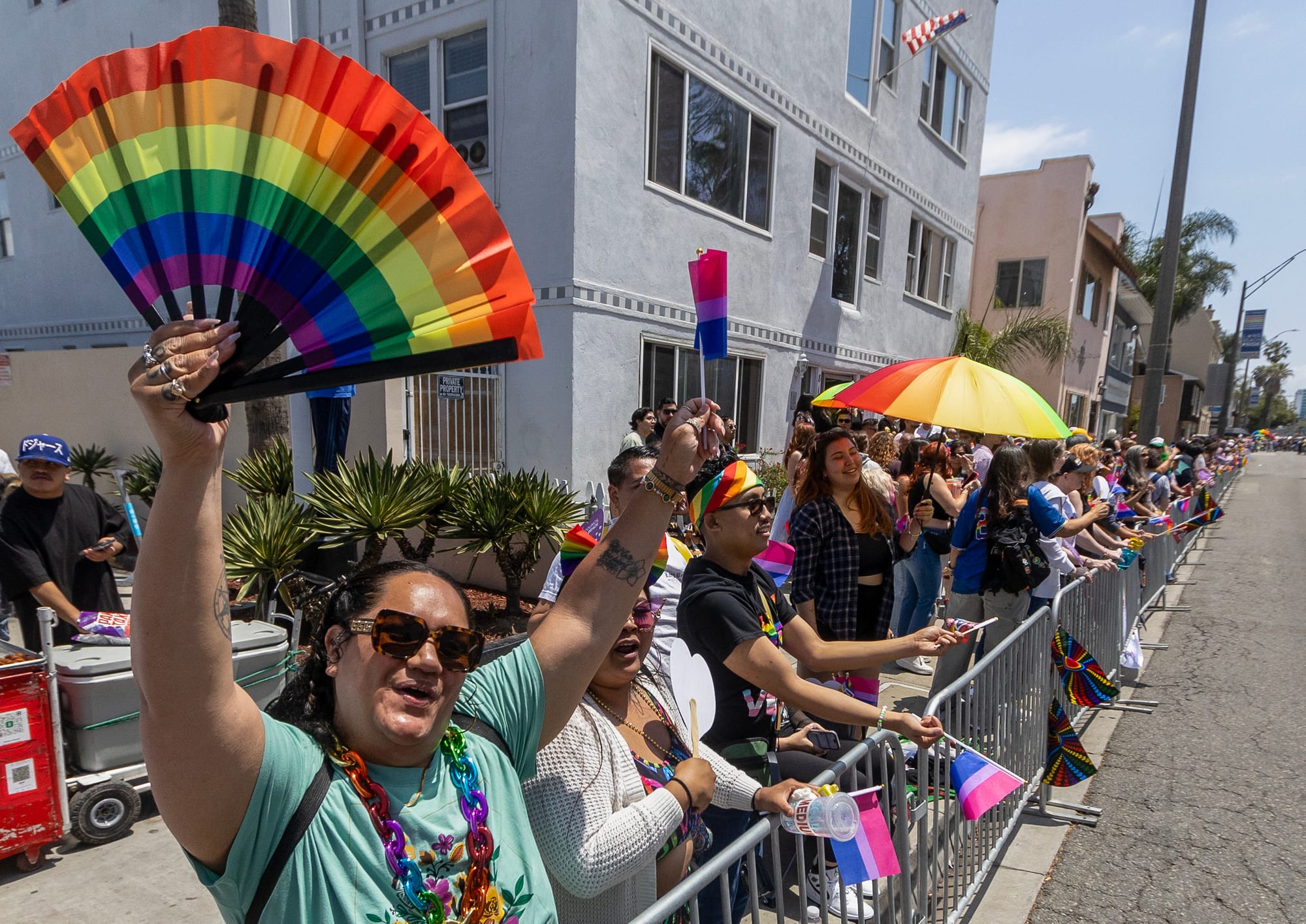 People stand behind a barricade on the sidewalk while waving colorful fans and flags.