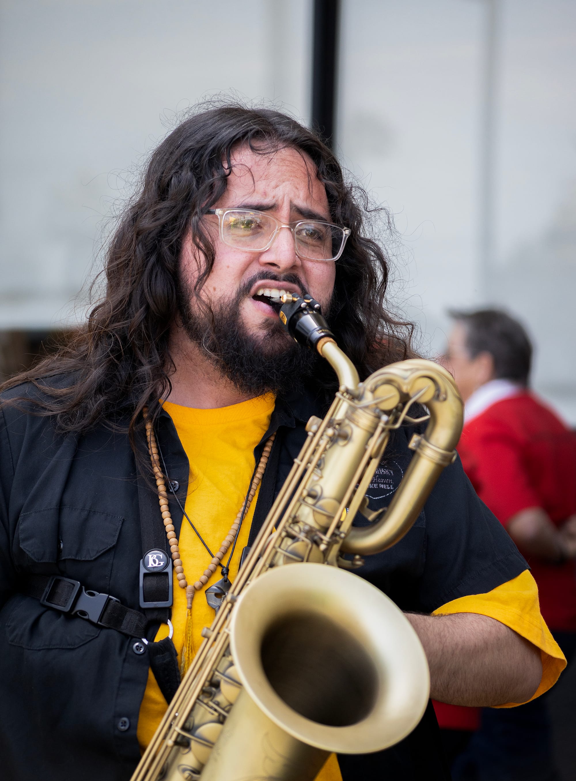 A man with long brown hair and glasses plays a saxophone.
