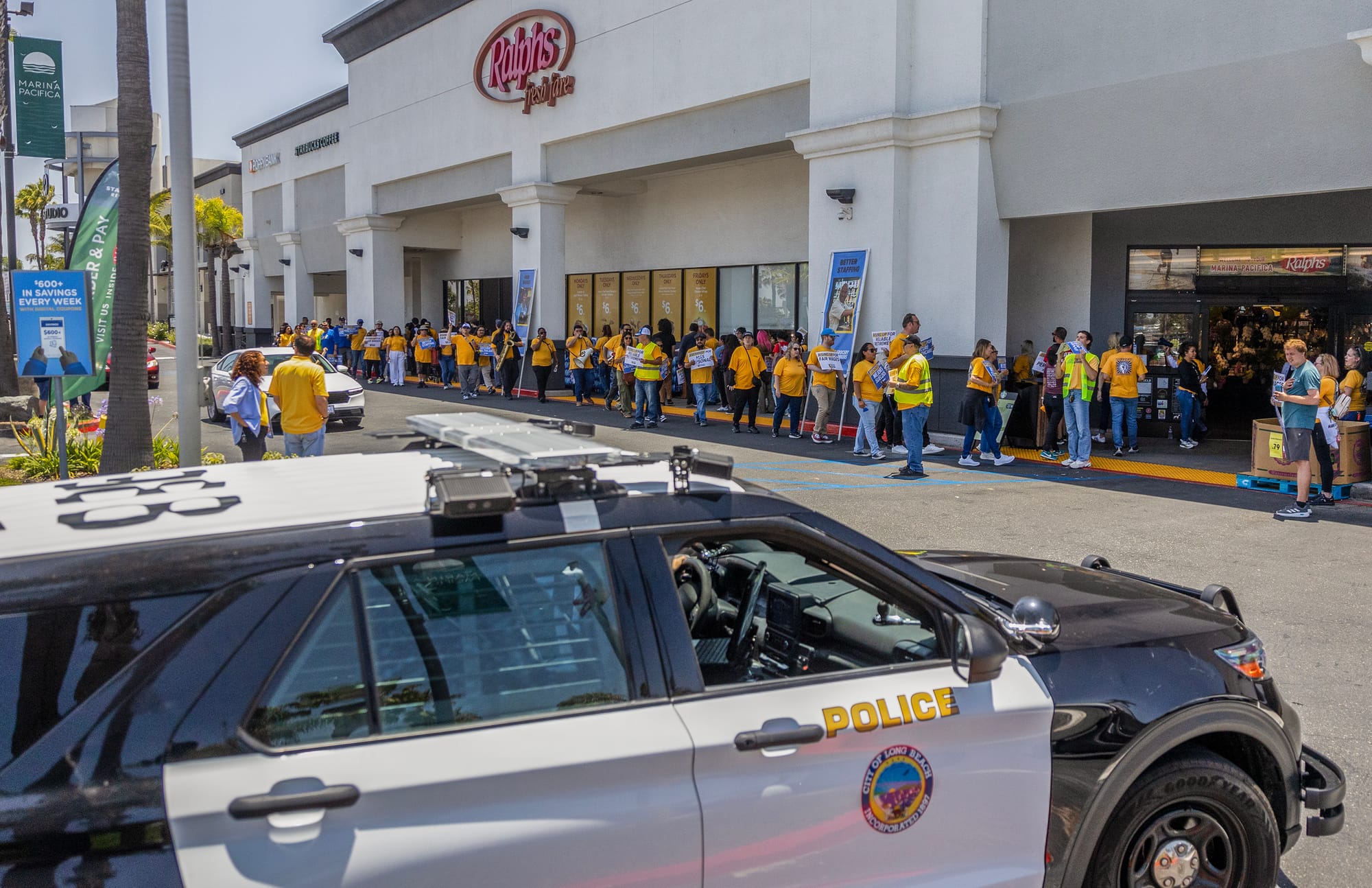 A police car is parked near a long line of people in yellow shirts standing outside a Ralphs grocery store.