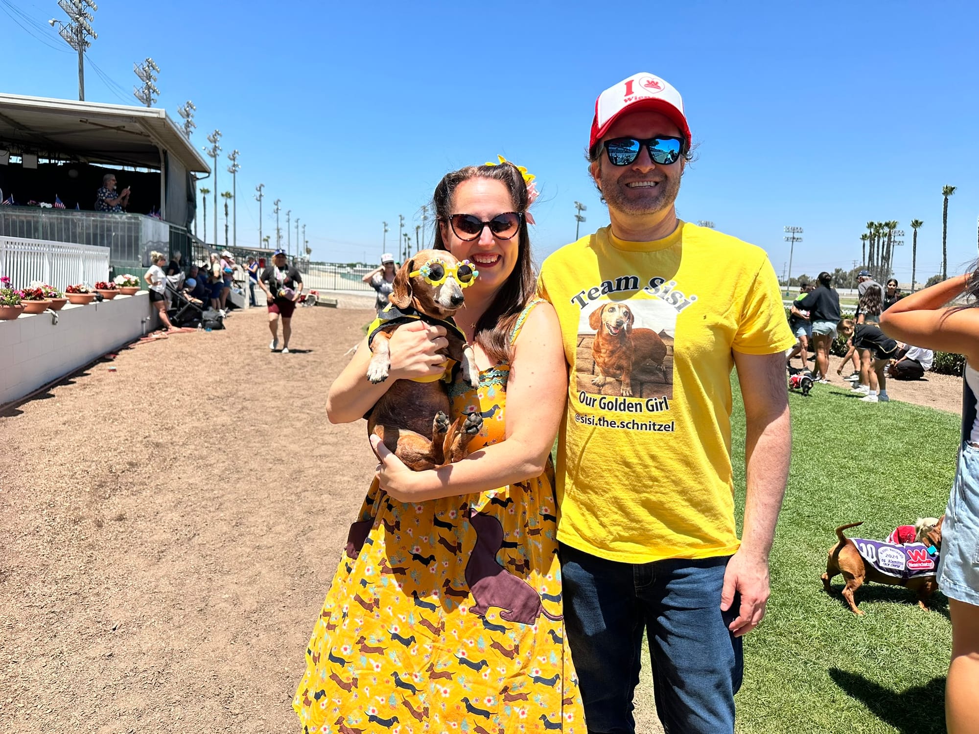 A smiling man stands next to a smiling woman holding a small brown dog wearing sunglasses.