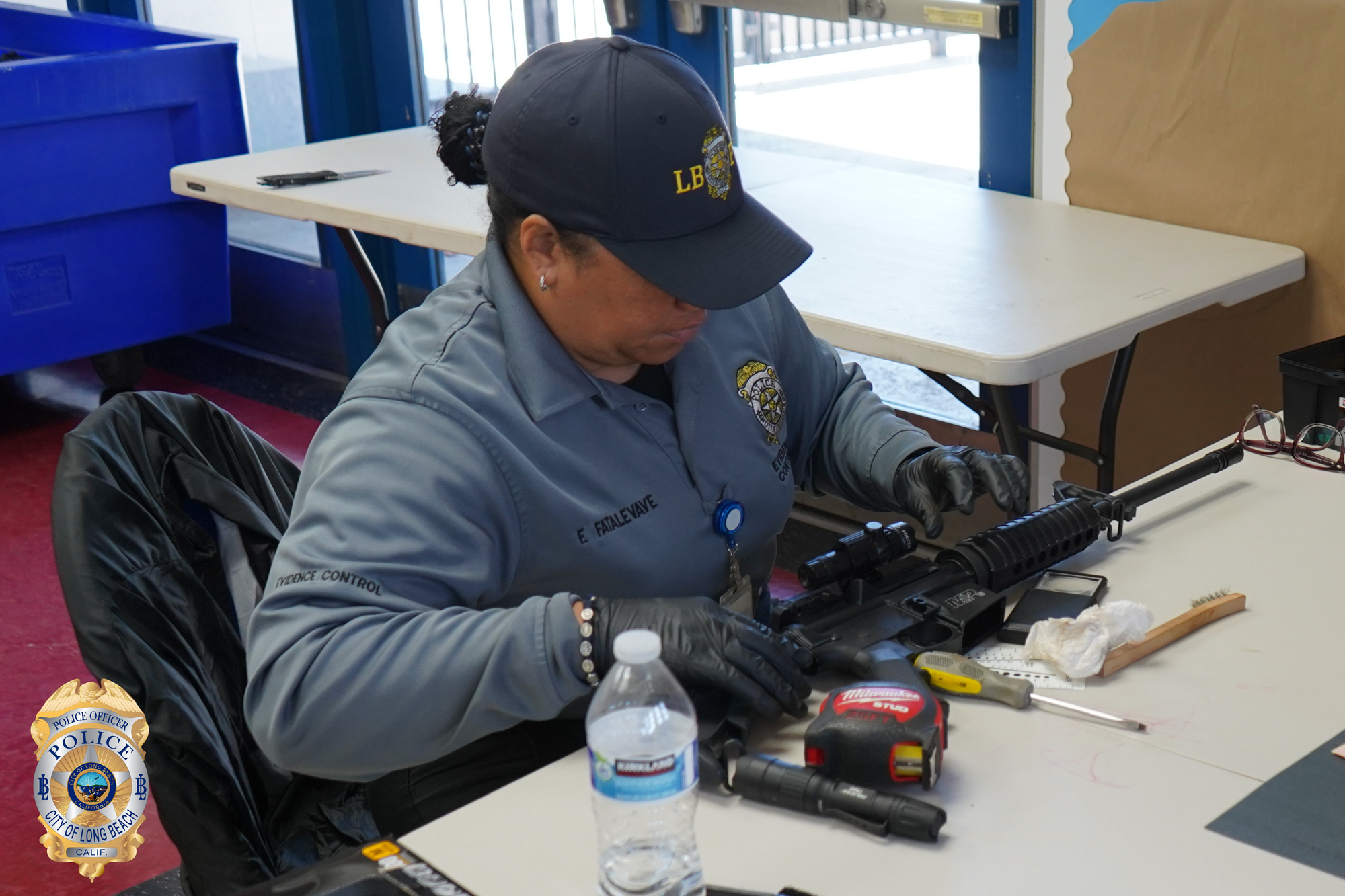 A person in a police polo shirt and baseball cap inspects a black rifle at a table.