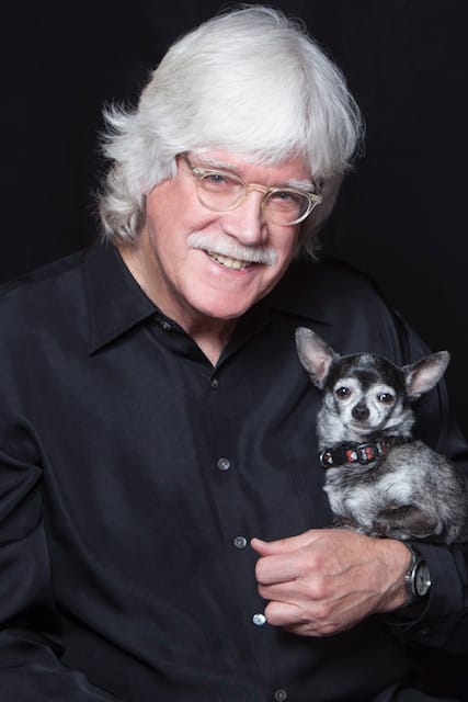 A man with white hair and glasses cradles a small black and gray dog.
