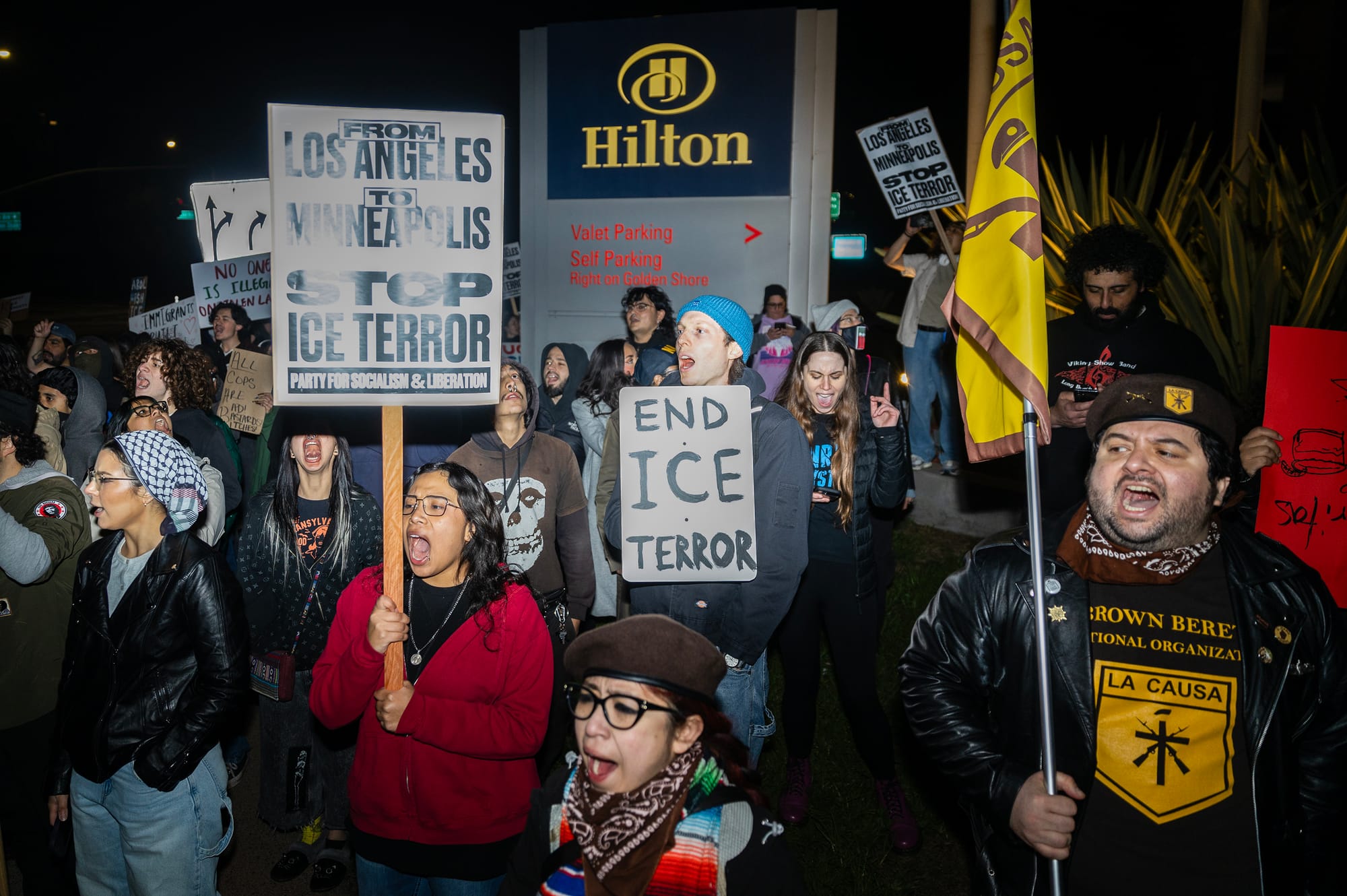 People chant and hold signs at night in front of a Hilton hotel sign.