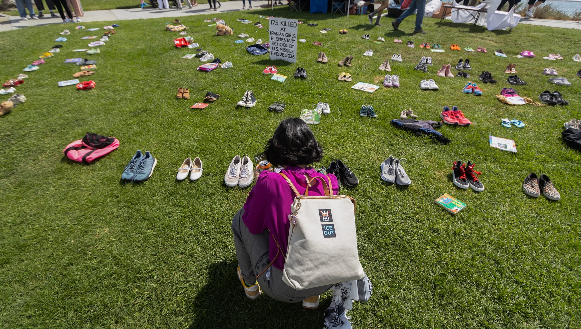 A woman with a canvas bag saying "Ice Out" kneels on the grass next to dozens of pairs of shoes.