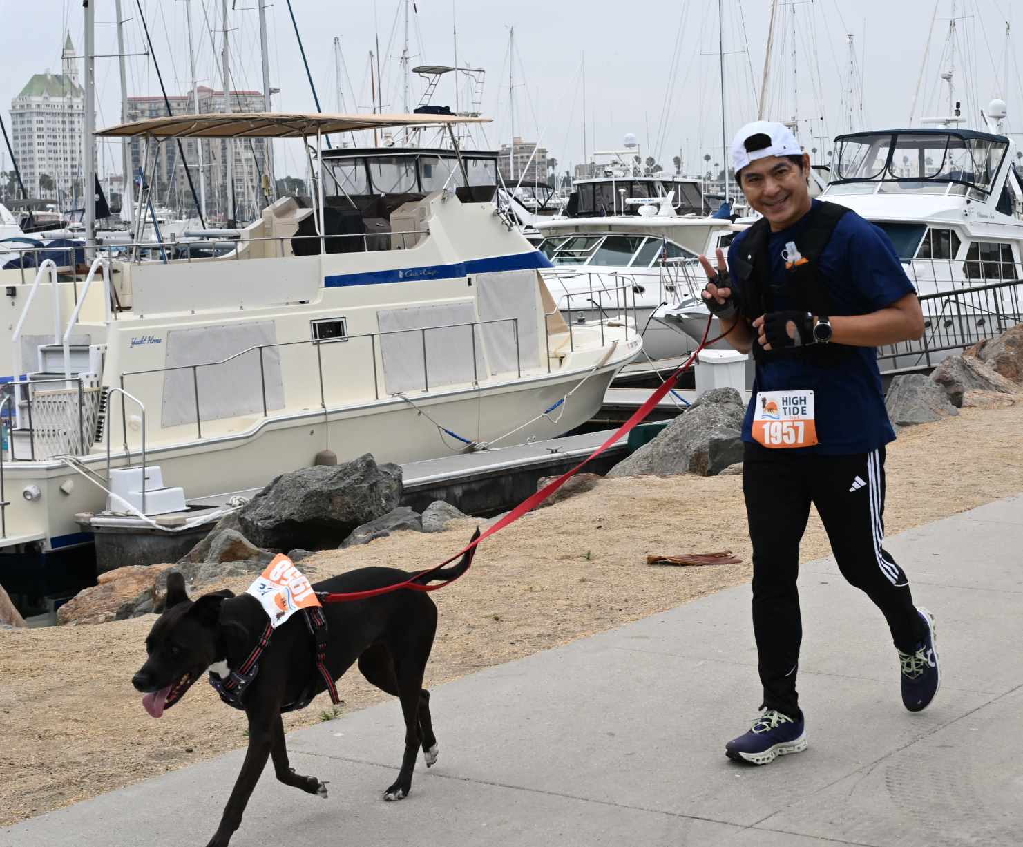 A man holds up two fingers while jogging with his dog next to a marina.