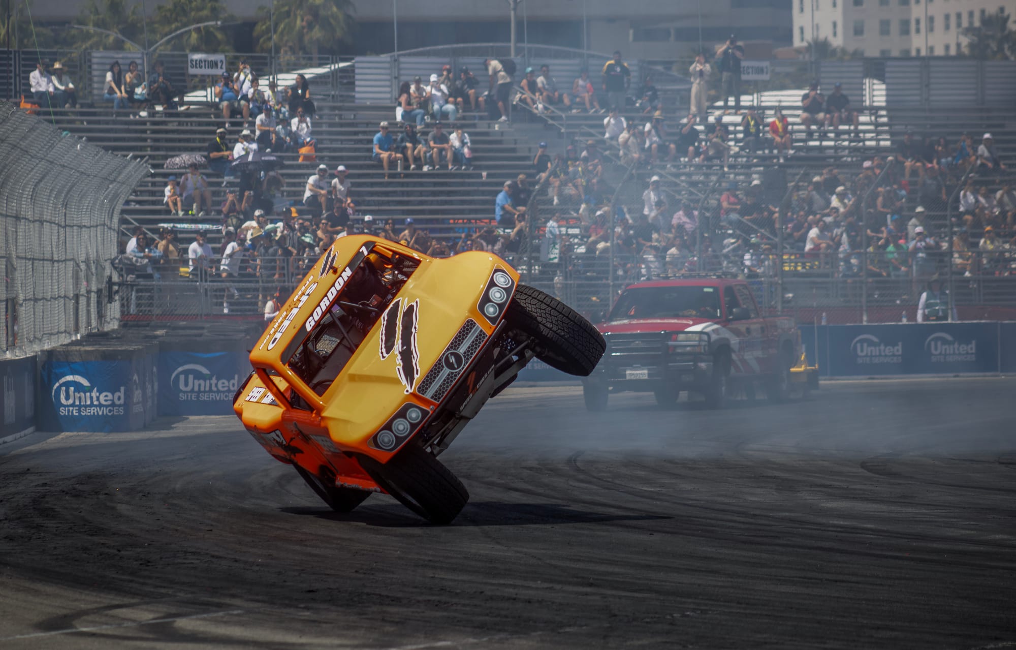 A yellow and orange truck rides on two wheels on a race track.