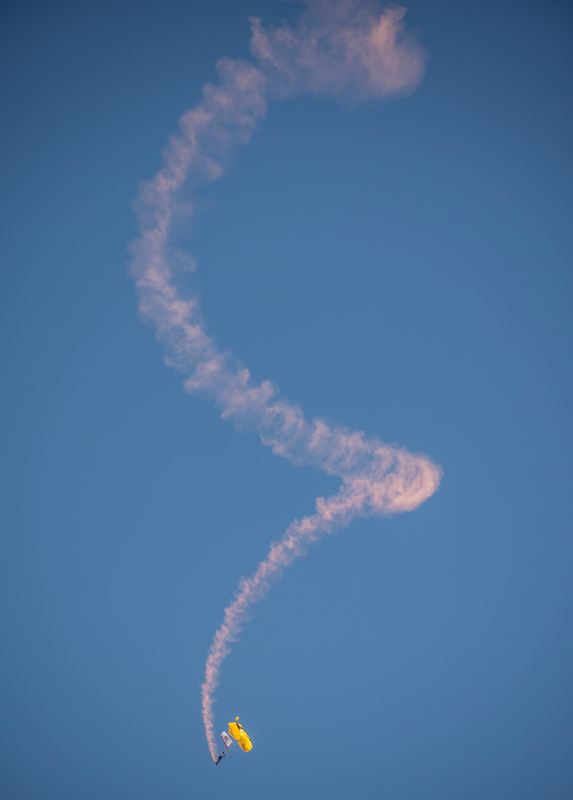 A parachutist leaves a wispy pink corkscrew of smoke on a clear blue sky.