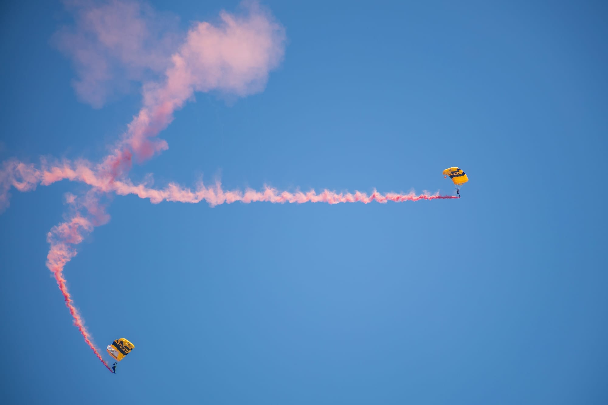 Two parachutists leave pink wispy smoke as they float down.