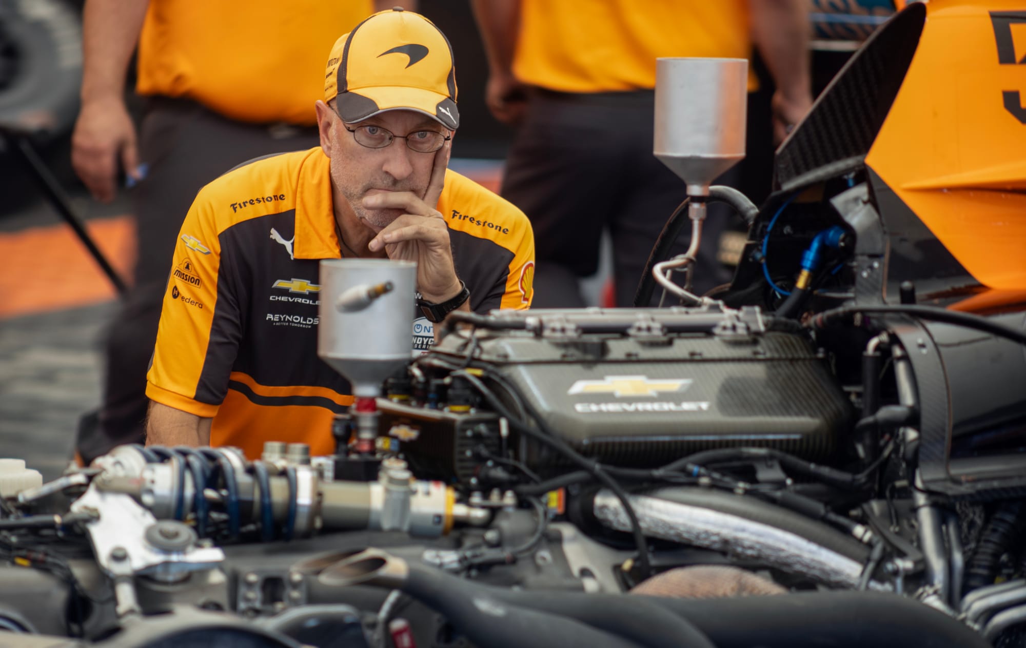A man wearing glasses and a yellow hat studies a race car engine.