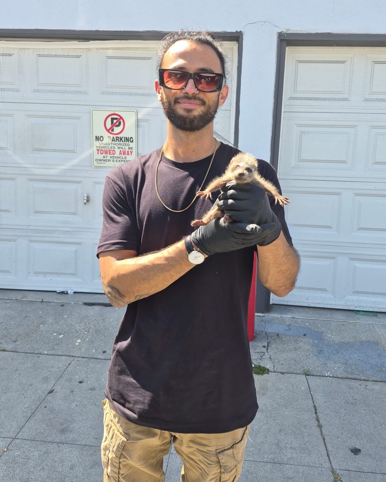 A man stands outside holding up a baby raccoon.