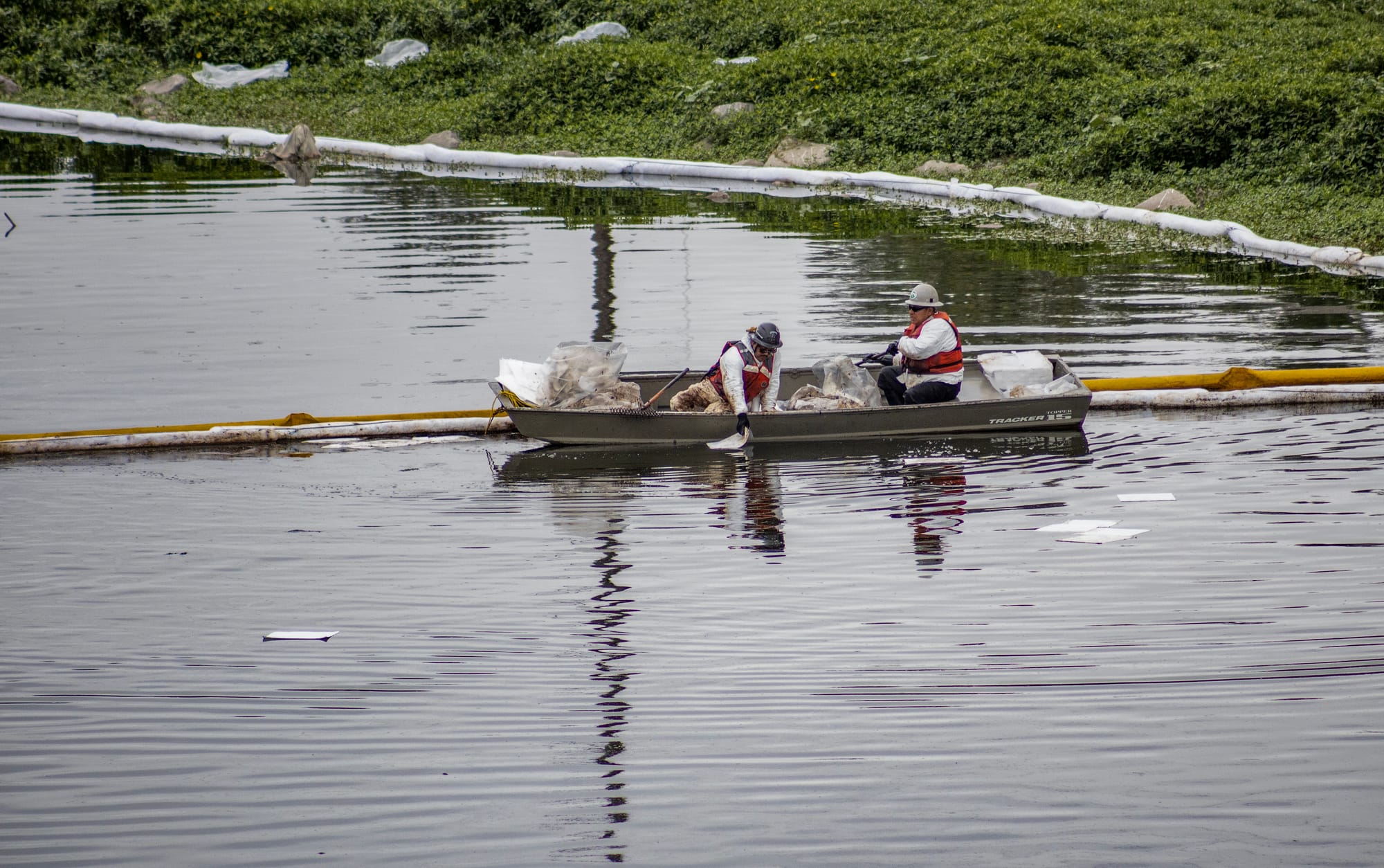 Two people in white coveralls and red life vests sit in a small gray boat on a river next to a long yellow tube.