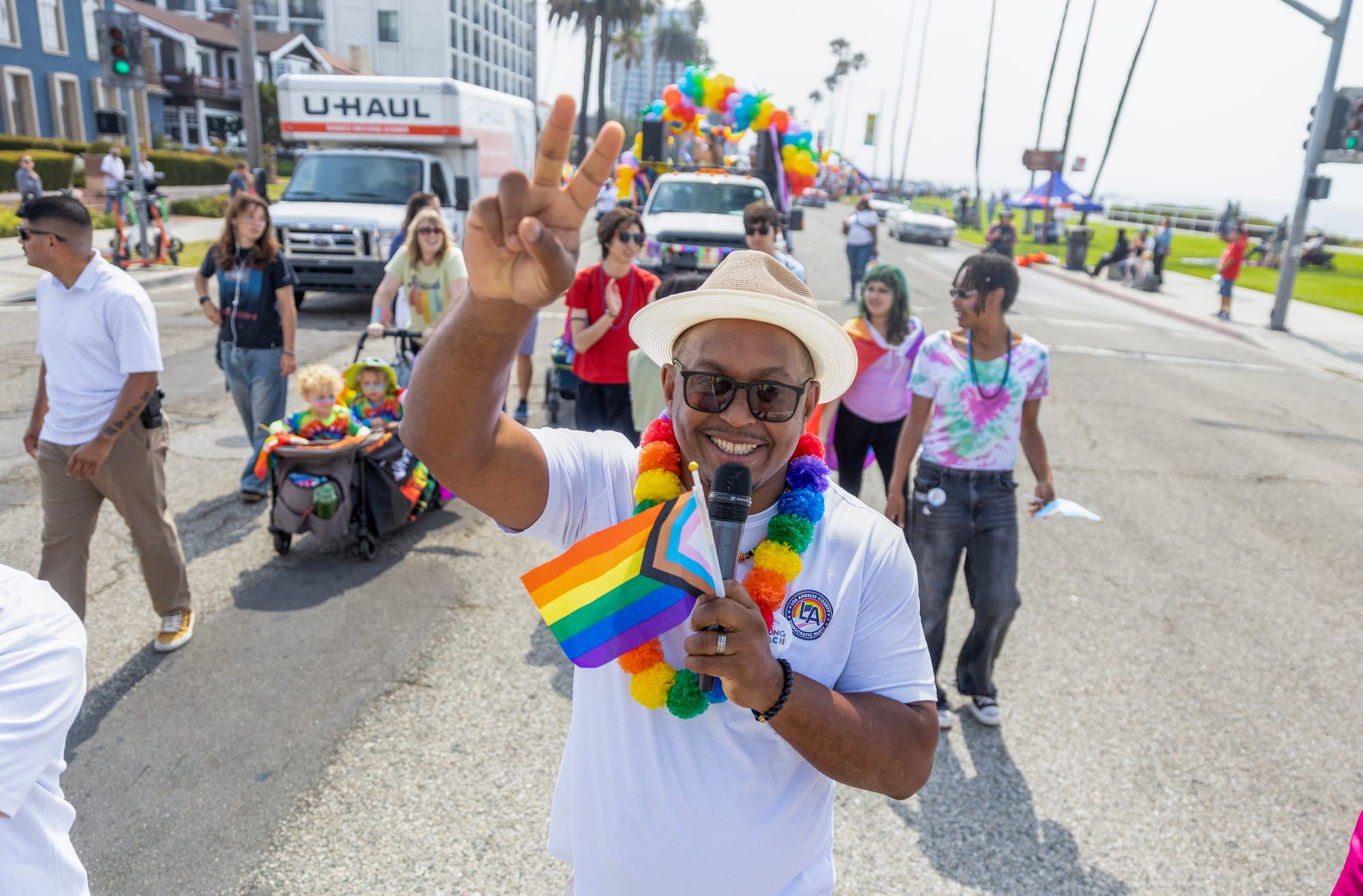 A man in a white shirt and white hat gives the peace sign with his fingers as he waves a tiny rainbow flag.