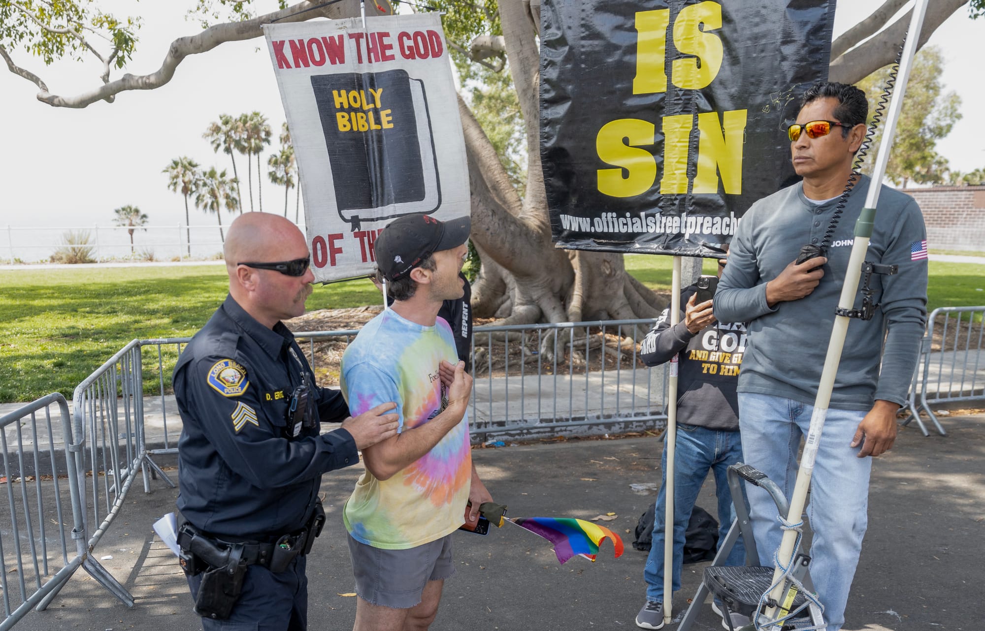 A police officer holds the shoulders of a man in a rainbow shirt whois yelling at a much taller man.