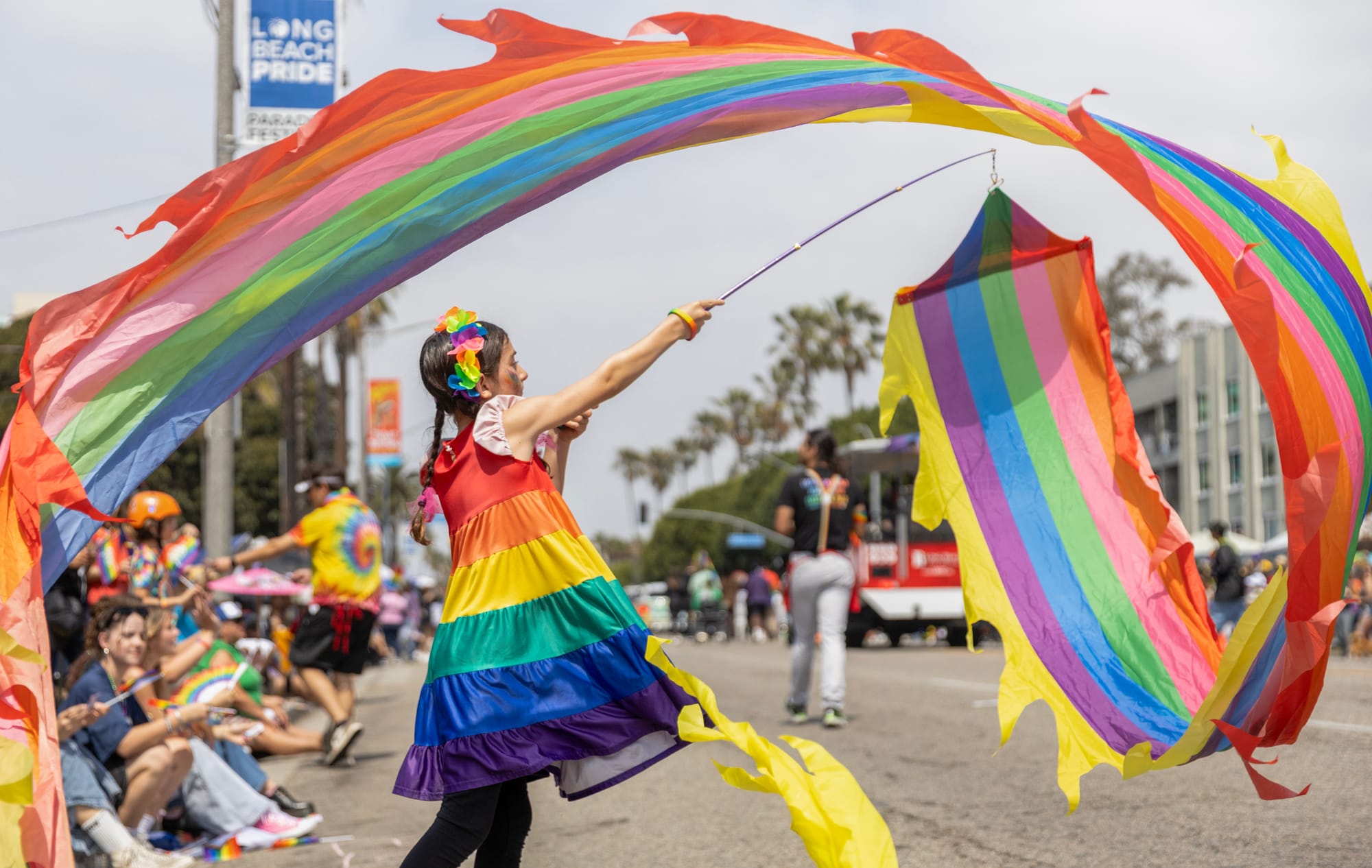 A little girl in a rainbow colored dress waves a long rainbow colored banner on the street.