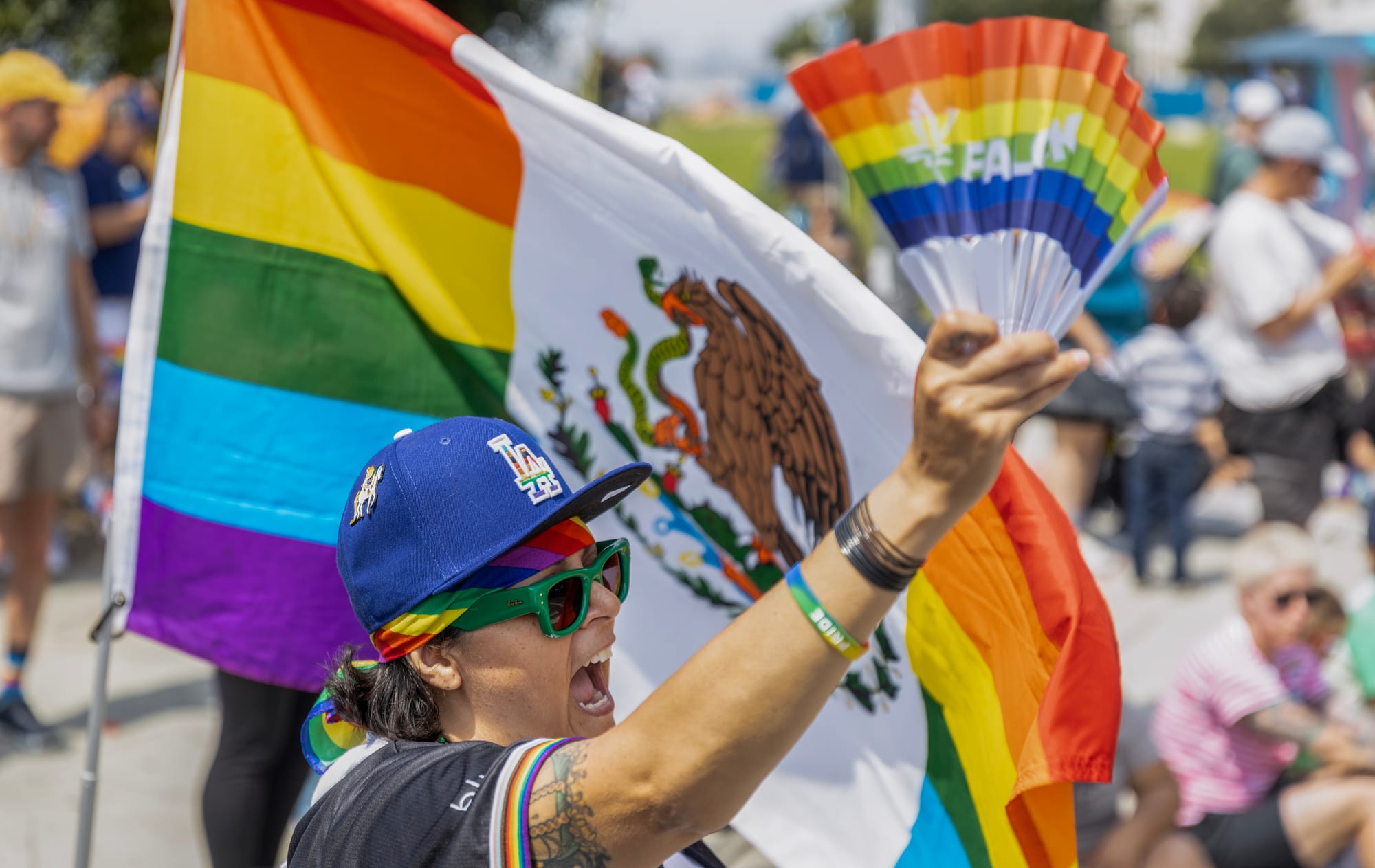 A person in a blue baseball cap and green sunglasses waves a small rainbow-colored fan in front of a large rainbow flag.