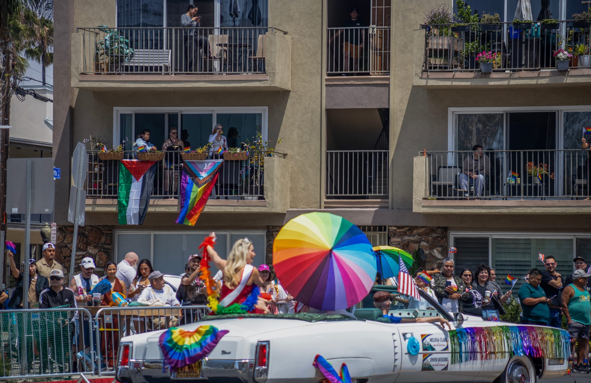 A large white convertible car with a giant rainbow umbrella in the back drives in front of a large group of people.