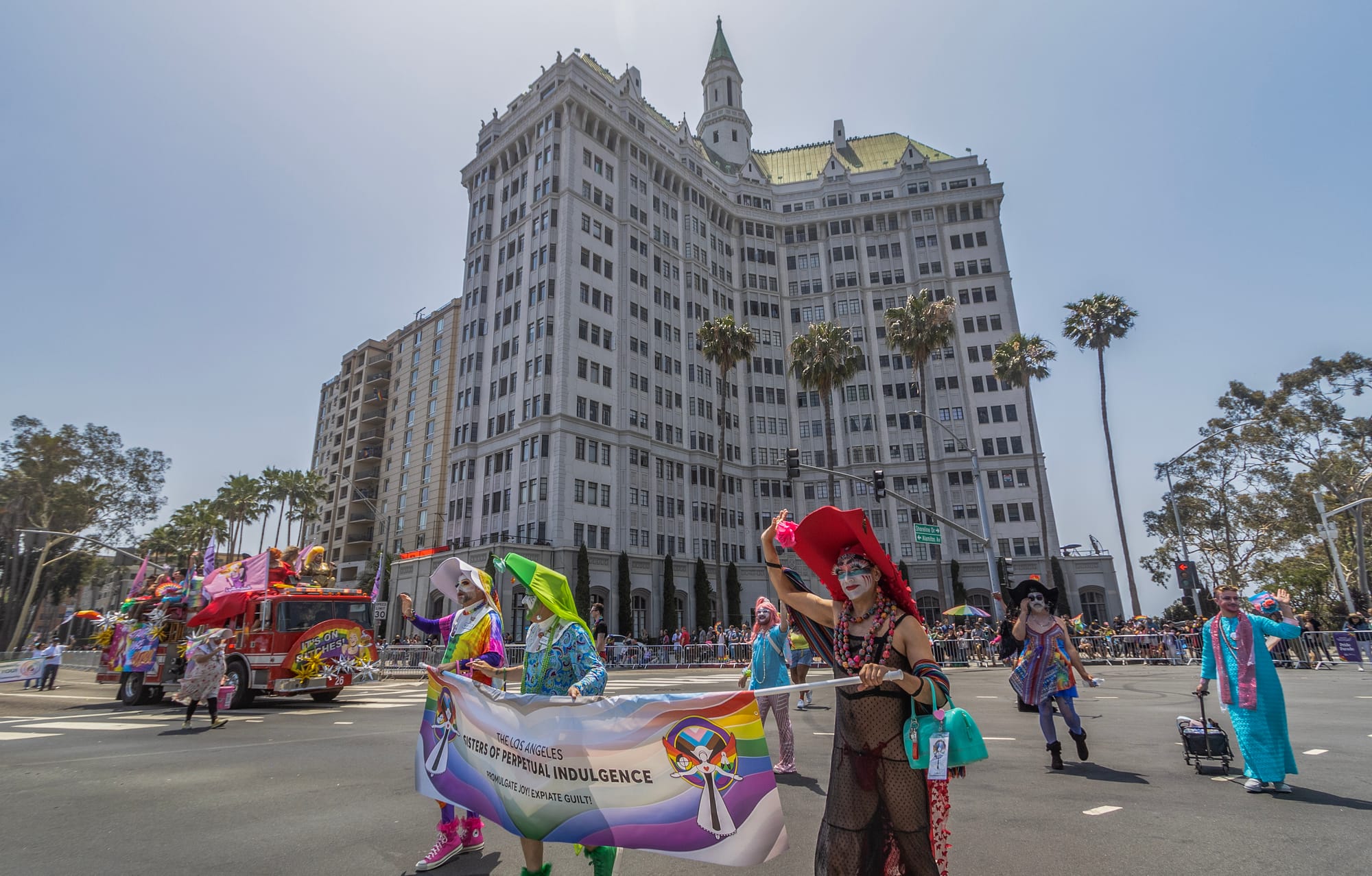 A small group of people in colorful costumes walk down the middle of a wide street in front of an old high-rise building.