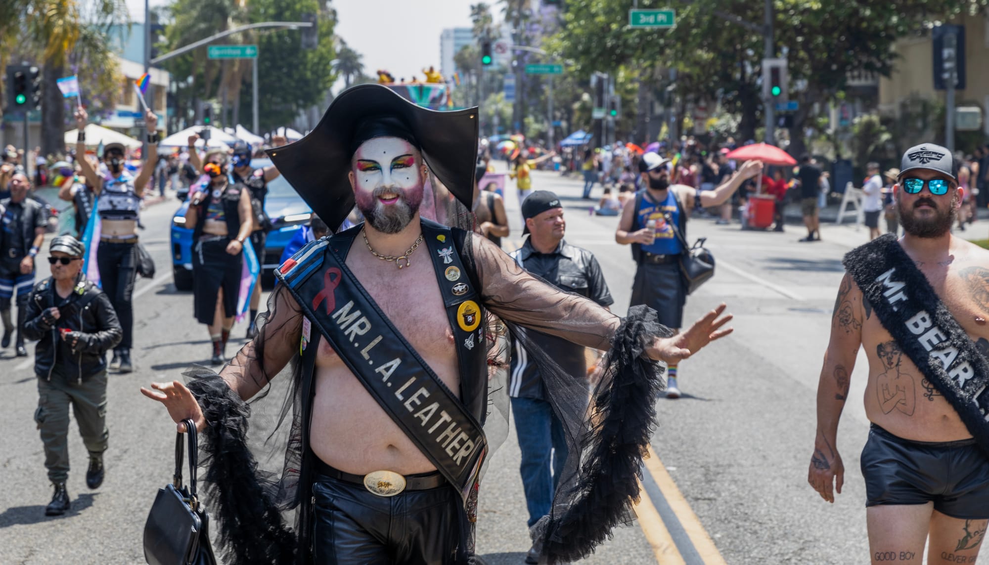 A person wears a black vest, nun's habit and sash saying "Mr. L. A. Leather" while walking in a parade.