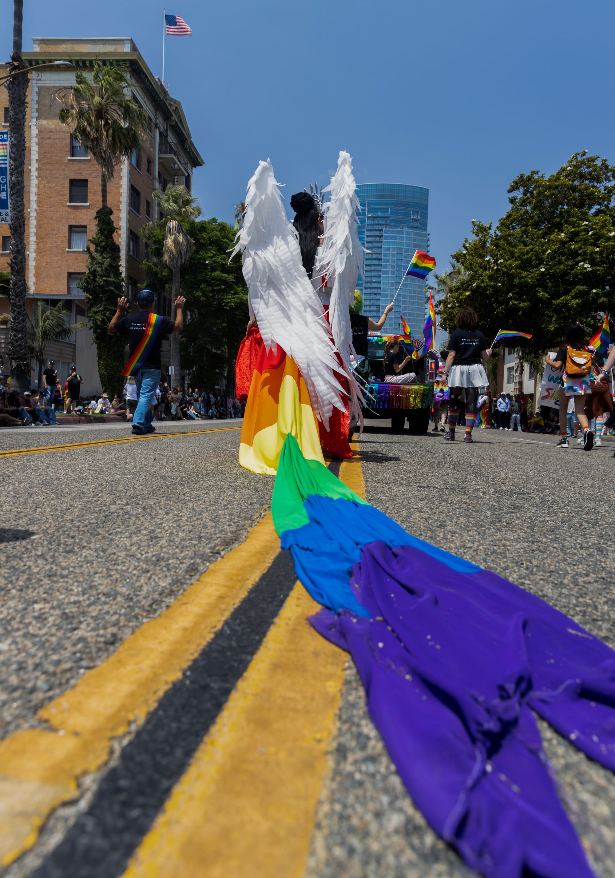 A long rainbow-colored train trails behind a person walking down the middle of the street.