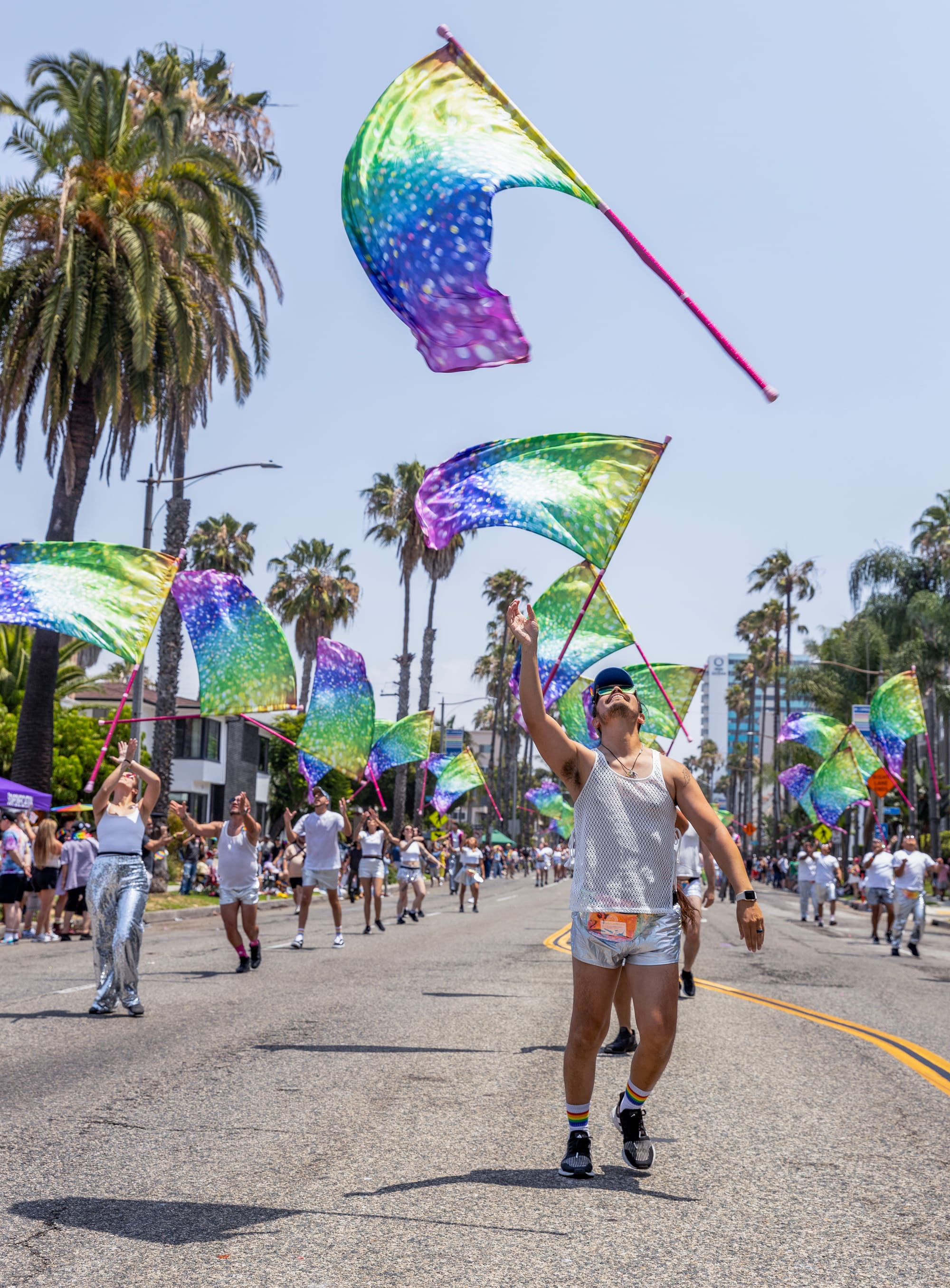 A man in a tanktop tosses a colorful flag into the air while walking down the street with other similarly dressed people.