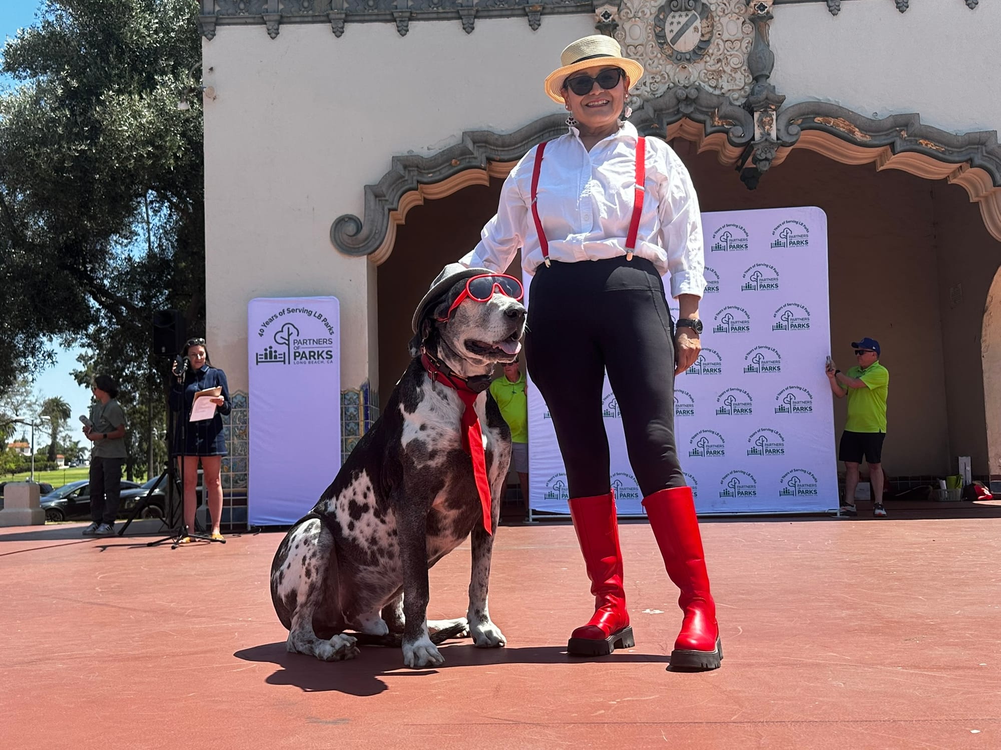 A woman stands next to a black and white dog wearing a hat, red tie and red sunglasses.