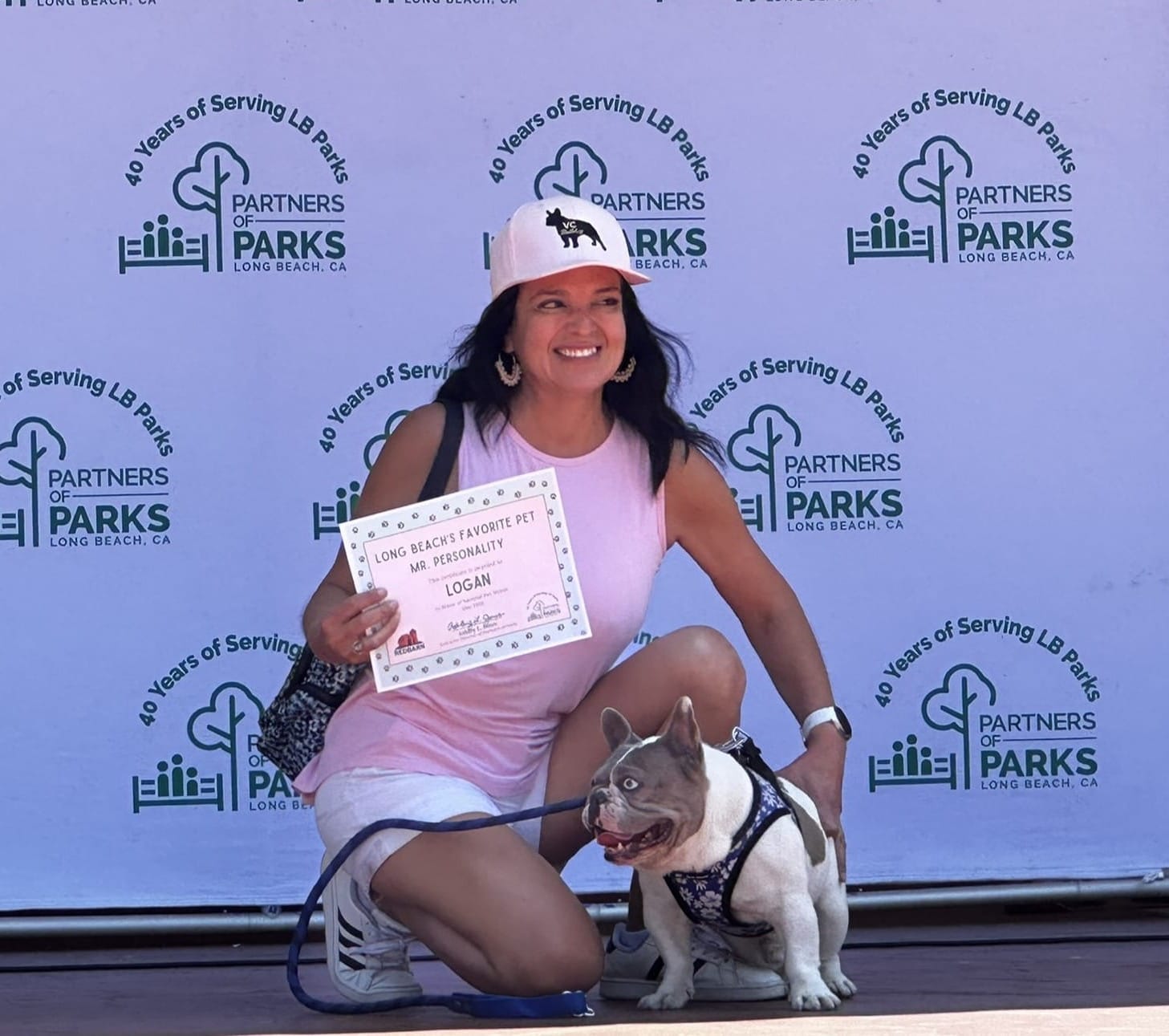 A woman holding an award certificate kneels on stage next to a small white and gray dog.