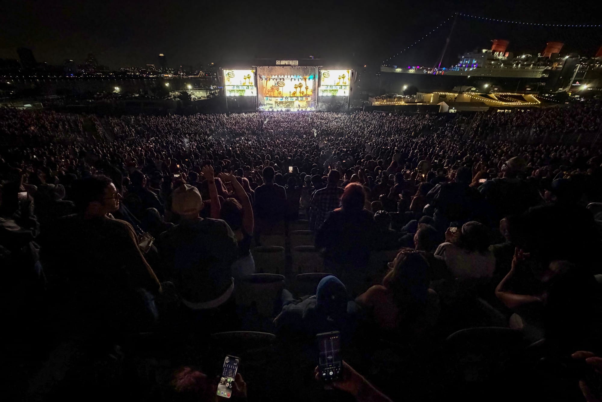 A large crowd of people sit in an arena at night next to a large brightly lit ship.