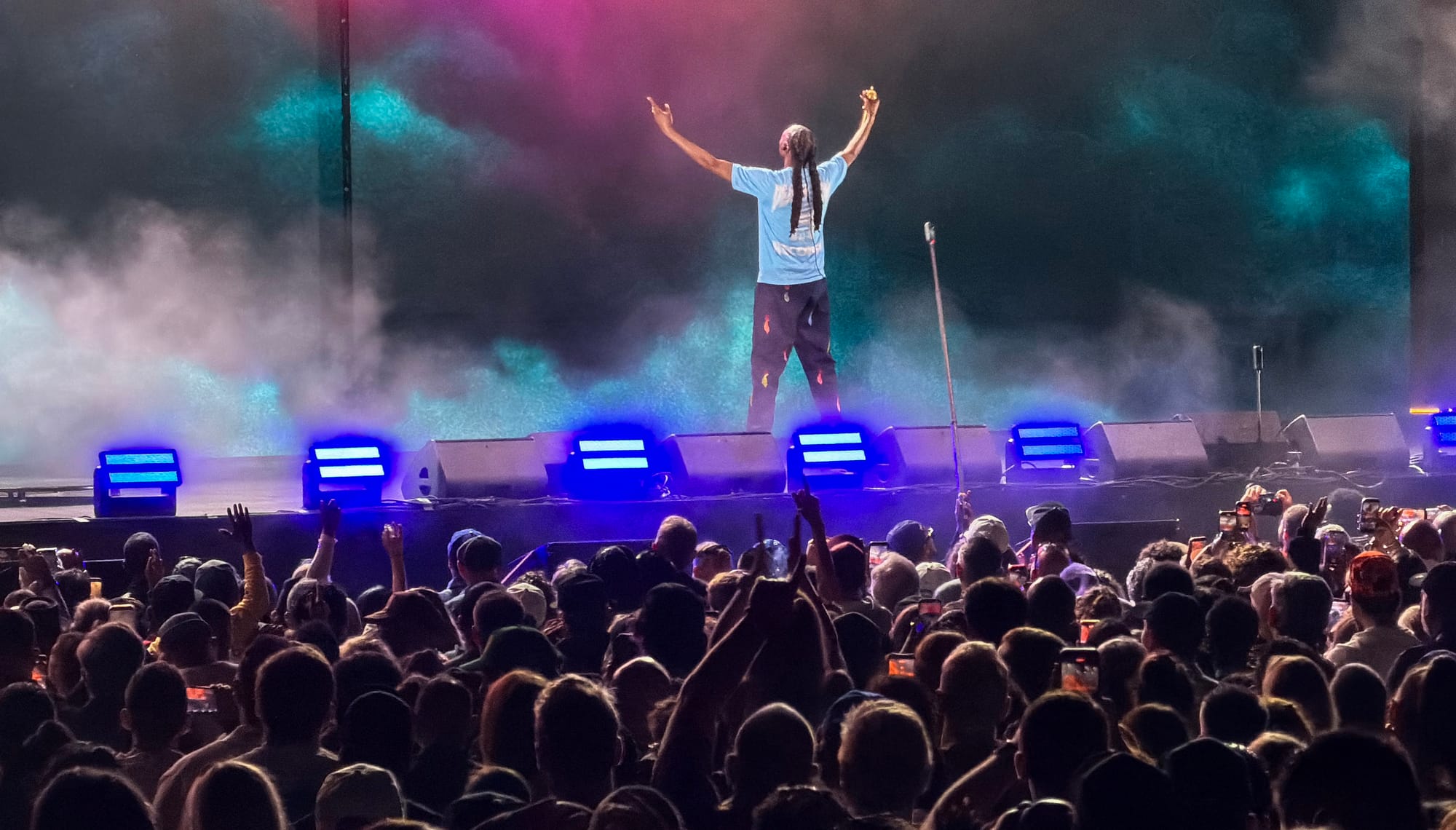 A man raises his arms high on stage in front of a large crowd at night.