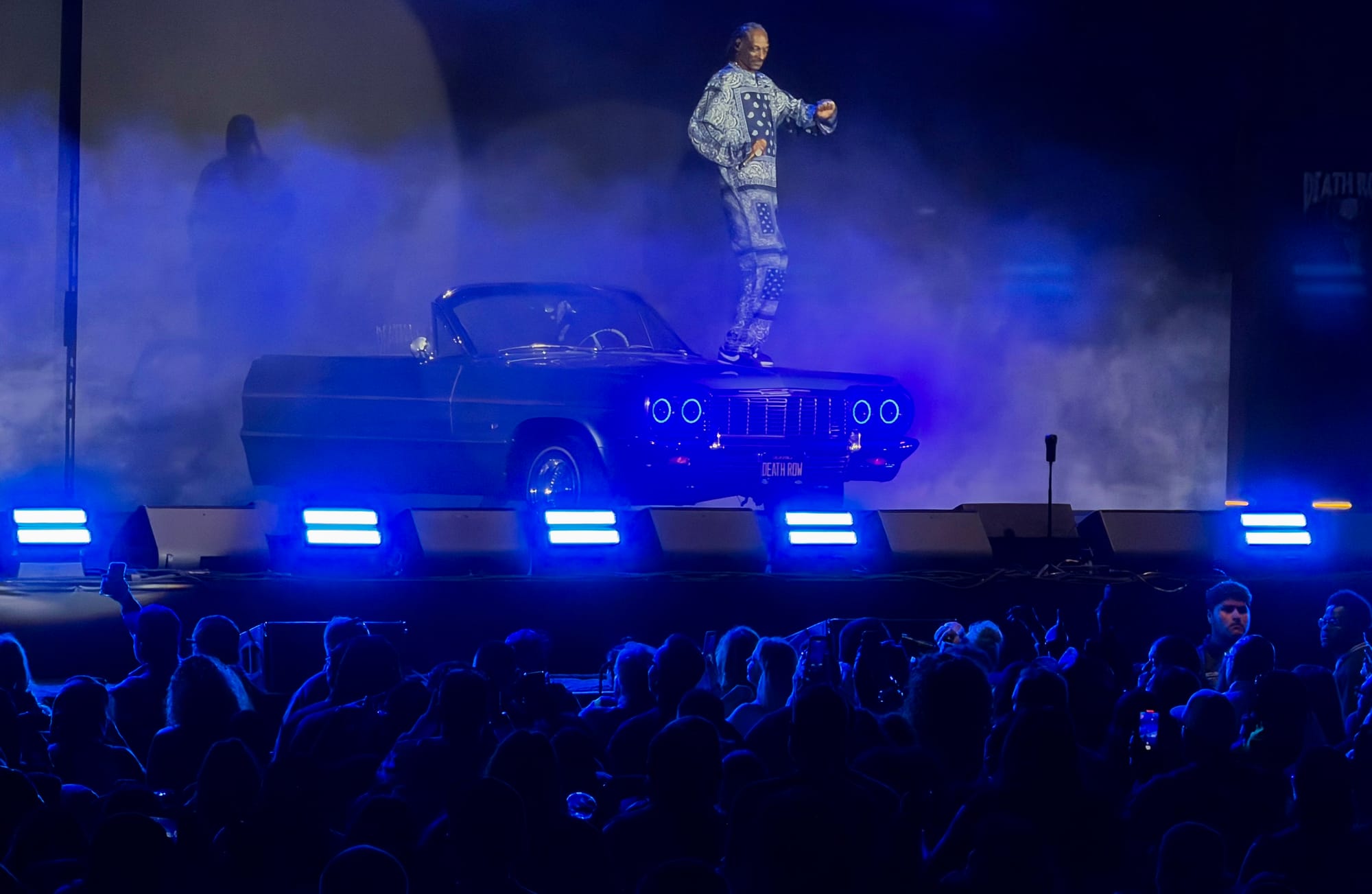 A man performs on the hood of a car onstage in front of a large crowd at night.