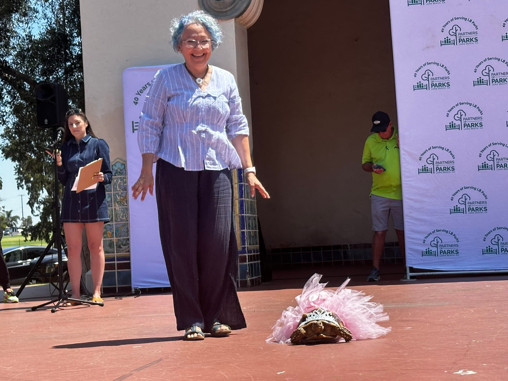 A smiling woman stands next to a turtle wearing a crown and pink tutu on a stage.