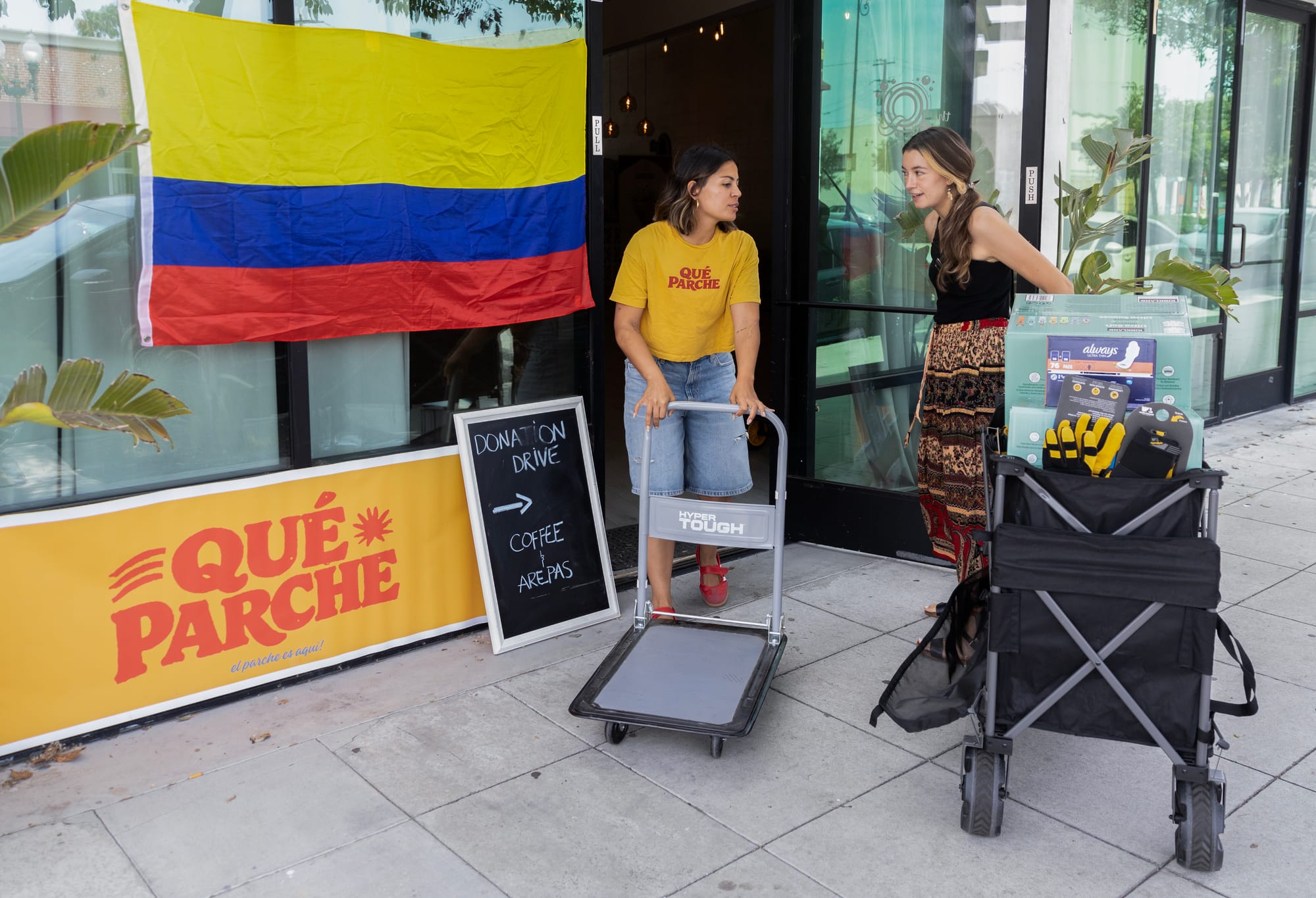 Two women stand on a sidewalk with carts next to a large flag with yellow, blue and red stripes.