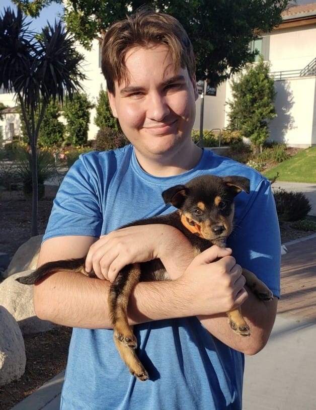 A man in a blue t-shirt cradles a little black and beige puppy.