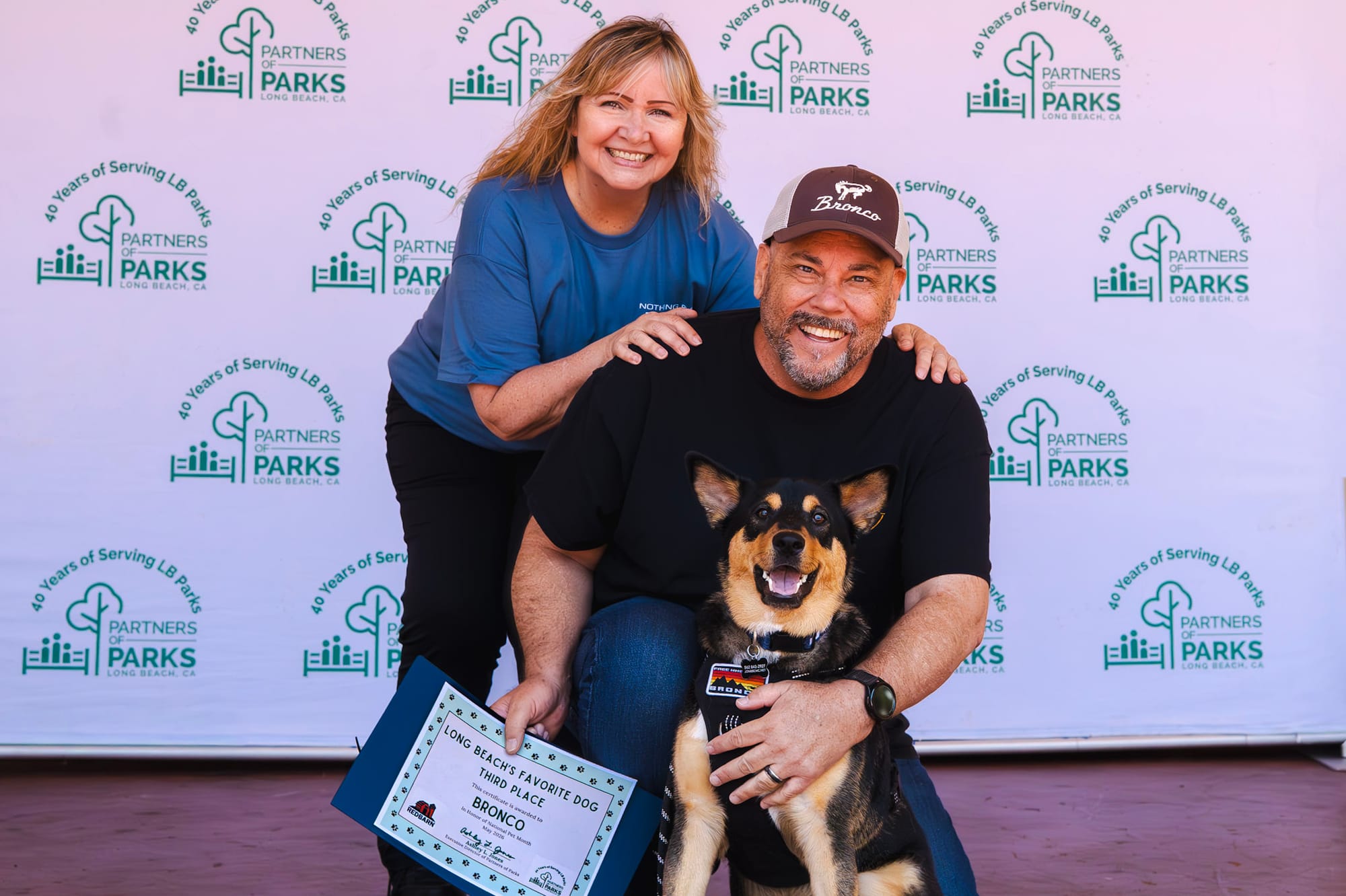 A man and woman smile while the man pets a happy black and beige dog.
