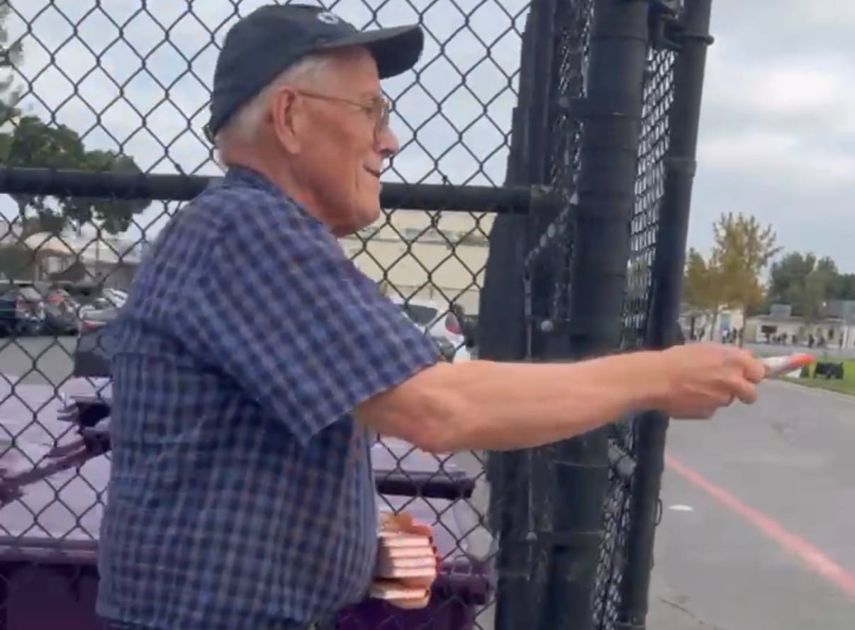 An old man holds out a book while standing near a fence.