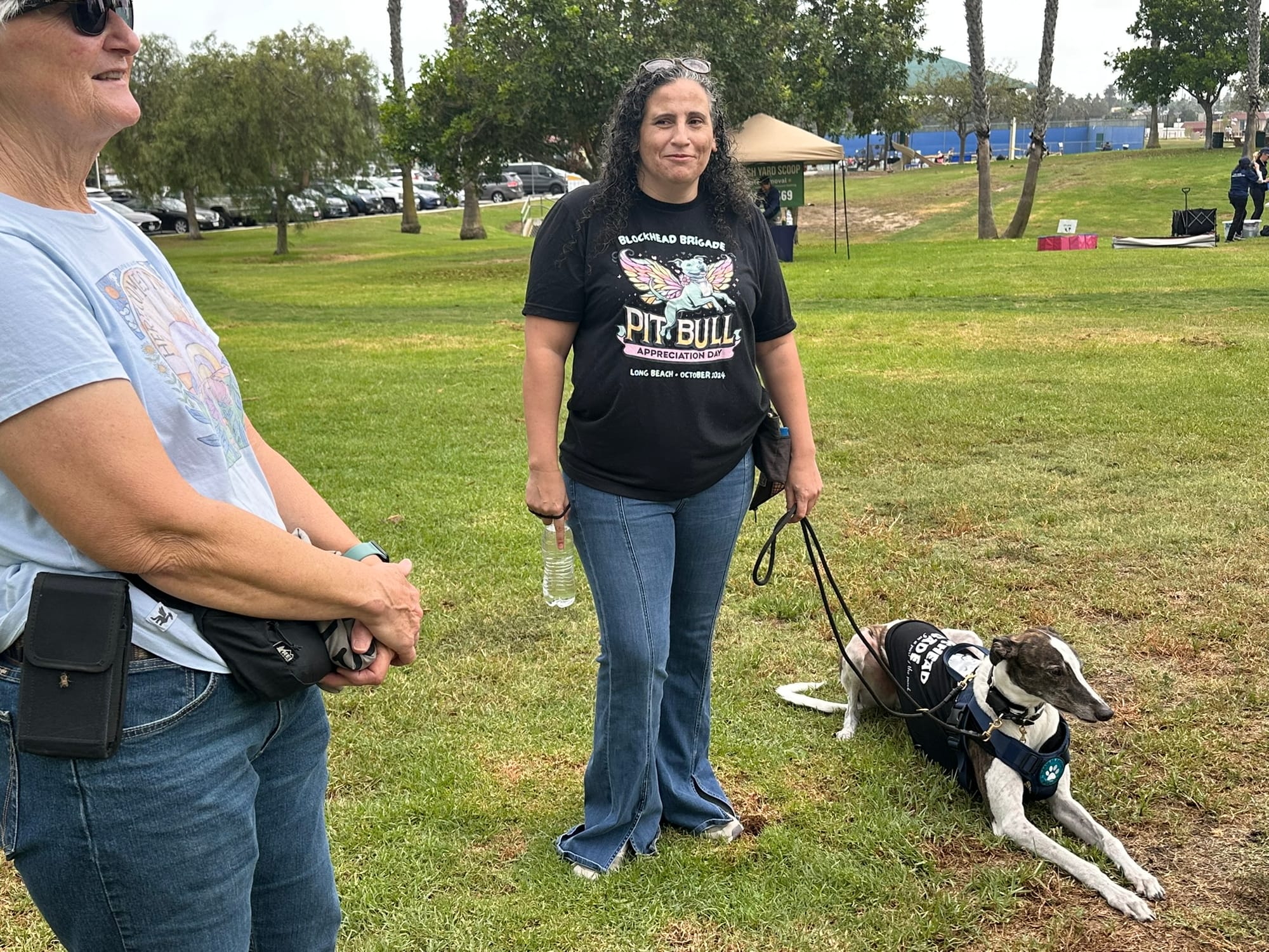 Two people stand next to a dog relaxing on the grass outside.