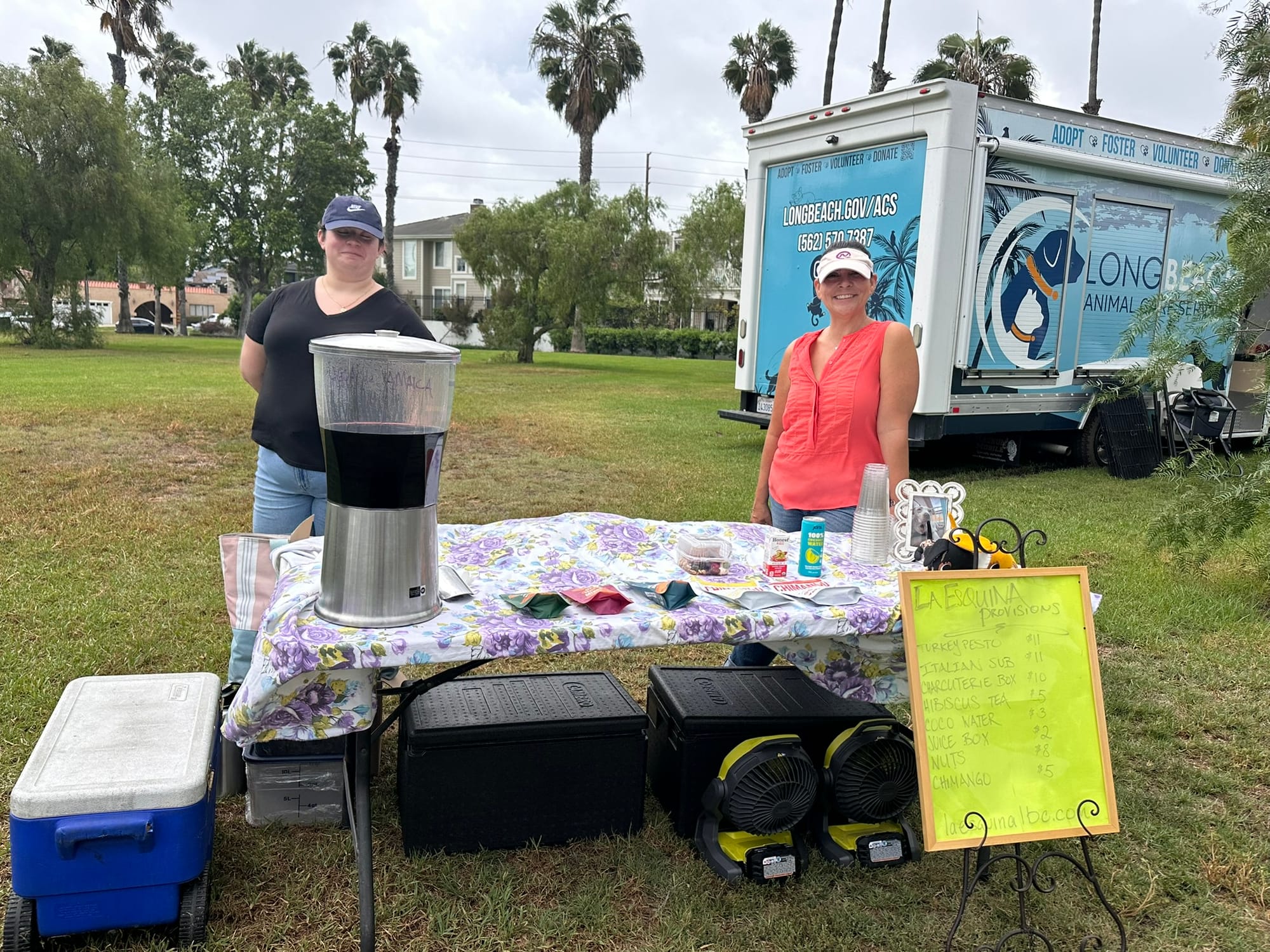 Two people stand behind a table covered in items near a large trailer.