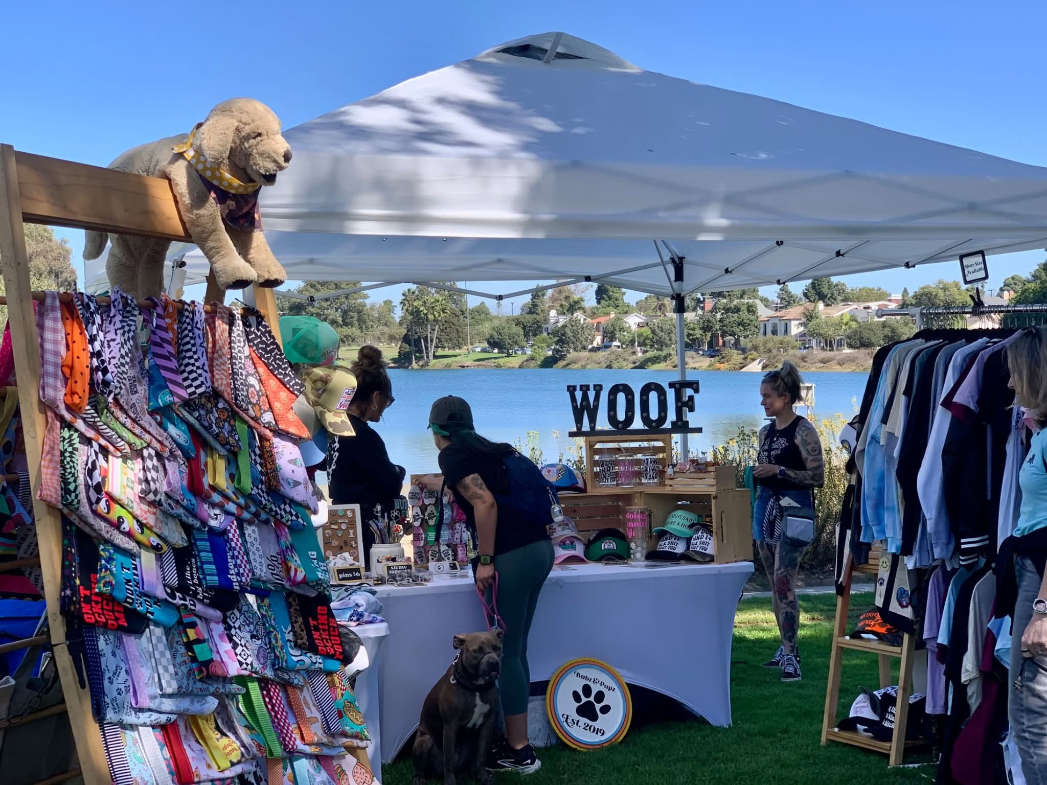 A person with a pit bull sitting near looks over items at a table under a white canopy.