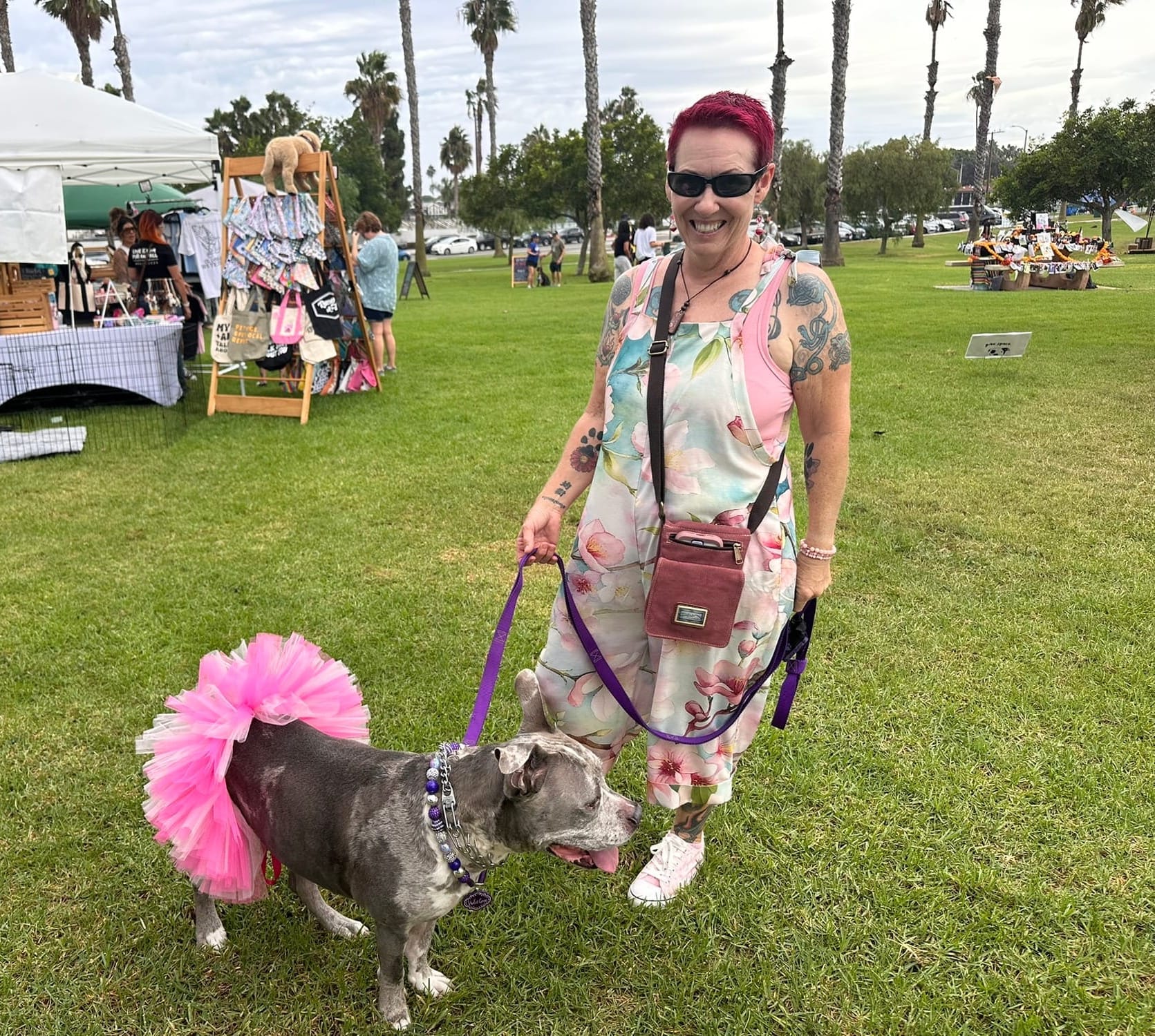 A smiling woman holds the purple leash of a dog wearing a pink tutu.