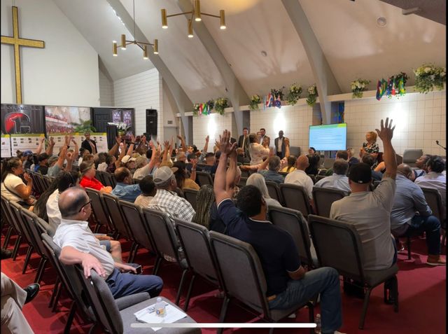 Dozens of people, many with their hands raised, sit in gray chairs in a large room.