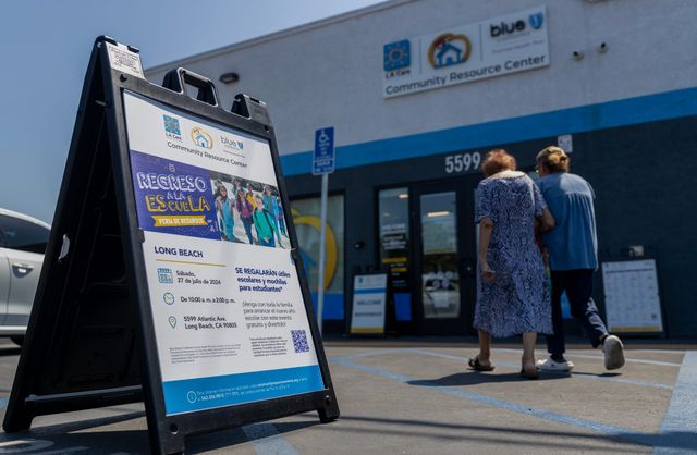 A white, purple and blue sign in Spanish announces an event while two elderly women walk toward a building.
