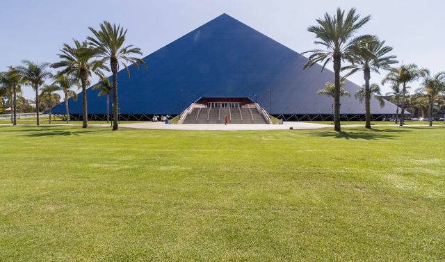 A big blue pyramid is surrounded by palm trees next to a green, grassy area.