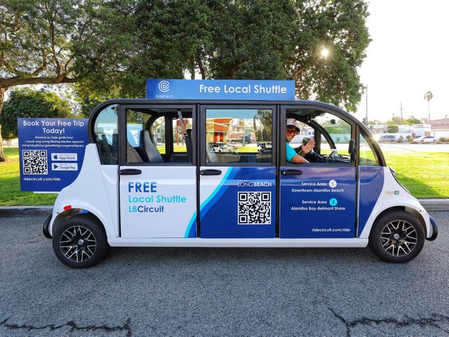 A person smiles in the driver's seat of a tiny white and blue micro-transit vehicle.