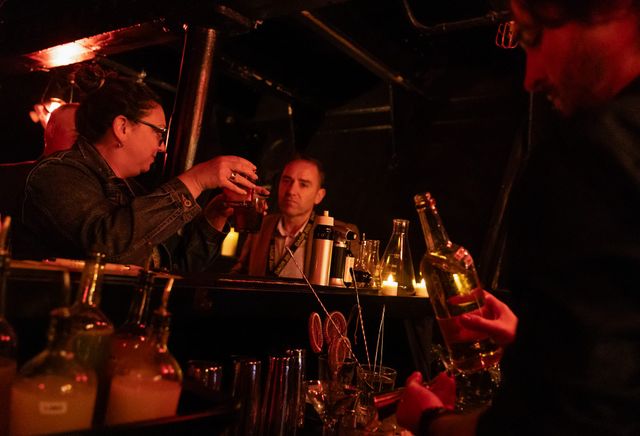 A man holds a bottle while two people clink glasses in a dark room.