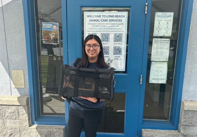 A woman holds a carrier with a cat inside while standing in front of top a door labeled "Welcome to Long Beach Animal Care Services."