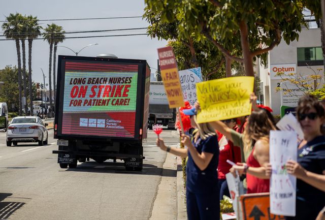 People stand on the street holding signs near a delivery van labeled "Long Beach nurses on strike for patient care!"