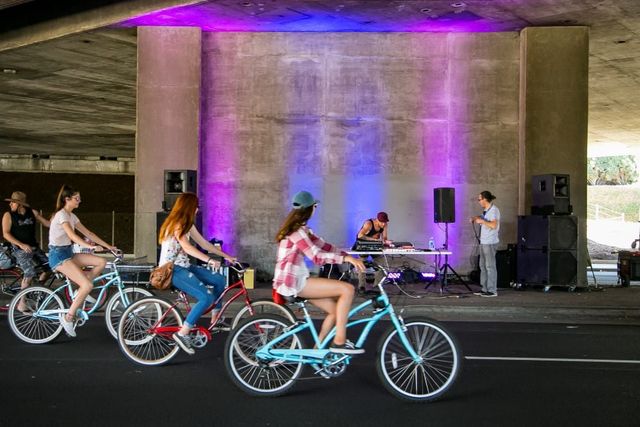 People ride bicycles past a man standing by a fold-up table.