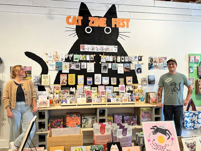 A man and woman stand on either side of a shelf covered in zines under the words "Cat Zine Fest."
