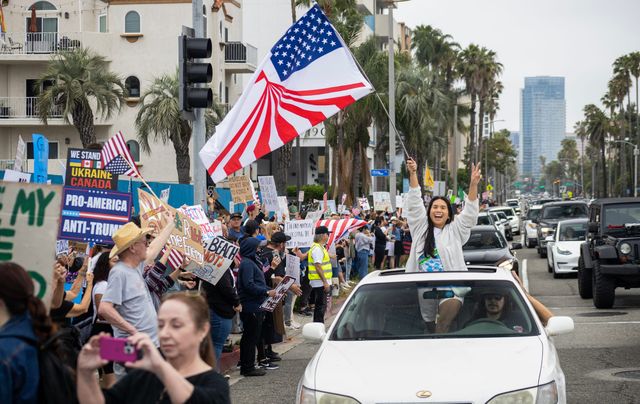 A woman smiles while standing in a car's sun roof and holding a stylized American flag.