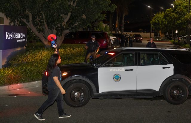 A man holding a bullhorn walks past a police car parked near a sign saying “Residence Inn.